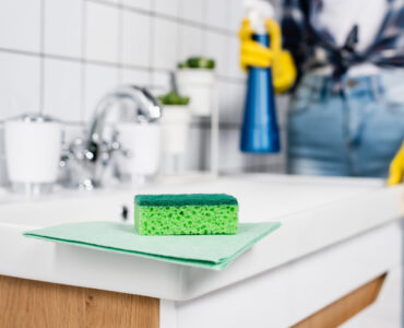 Green rag and sponge on sink near woman in rubber gloves cleaning bathroom on blurred background