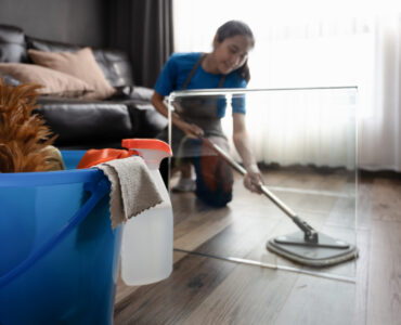 Young woman in an apron wiping laminate flooring in the living room with microfiber wet mop.